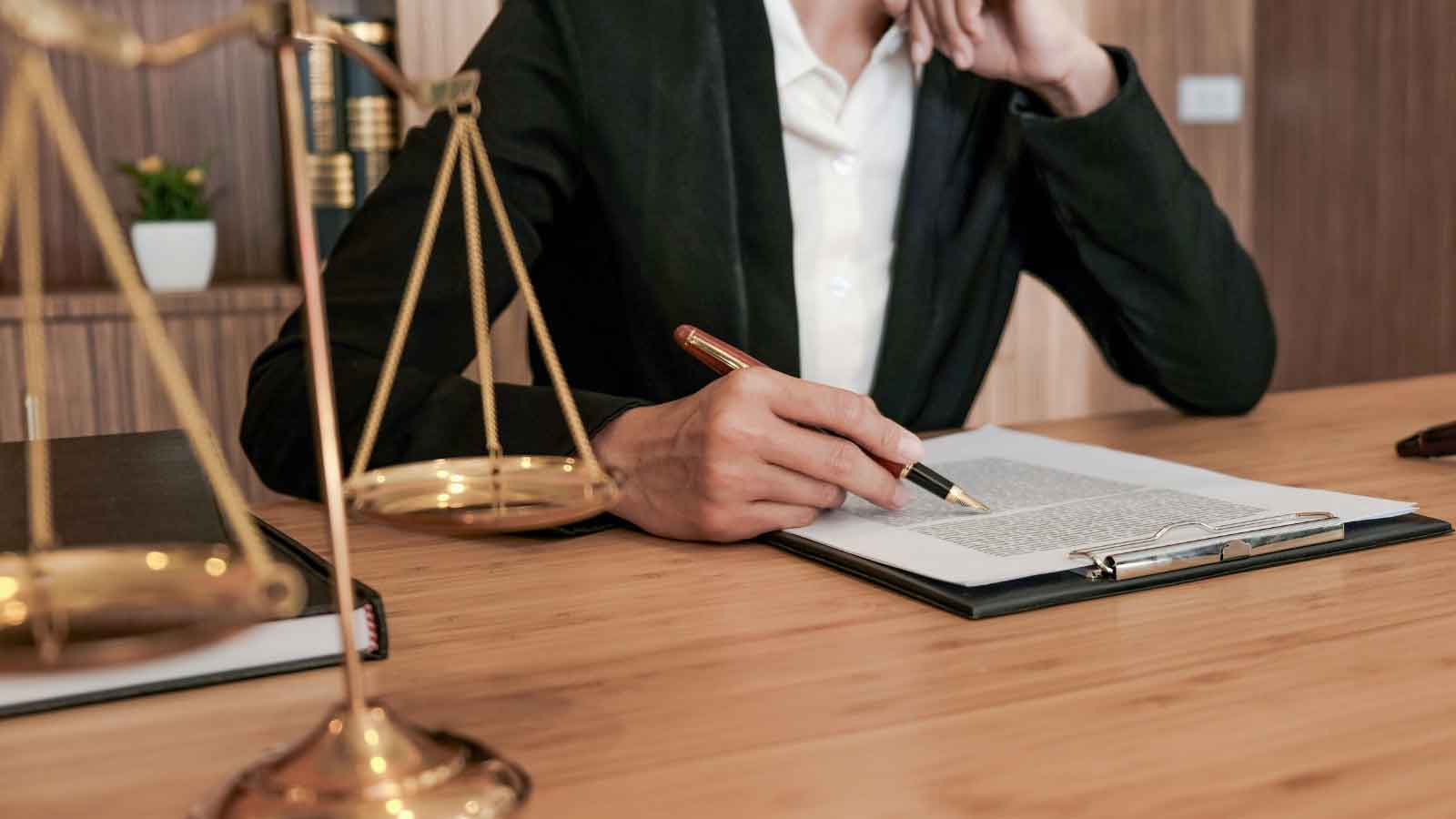 A person in a suit writing on a document with scales of justice in the foreground