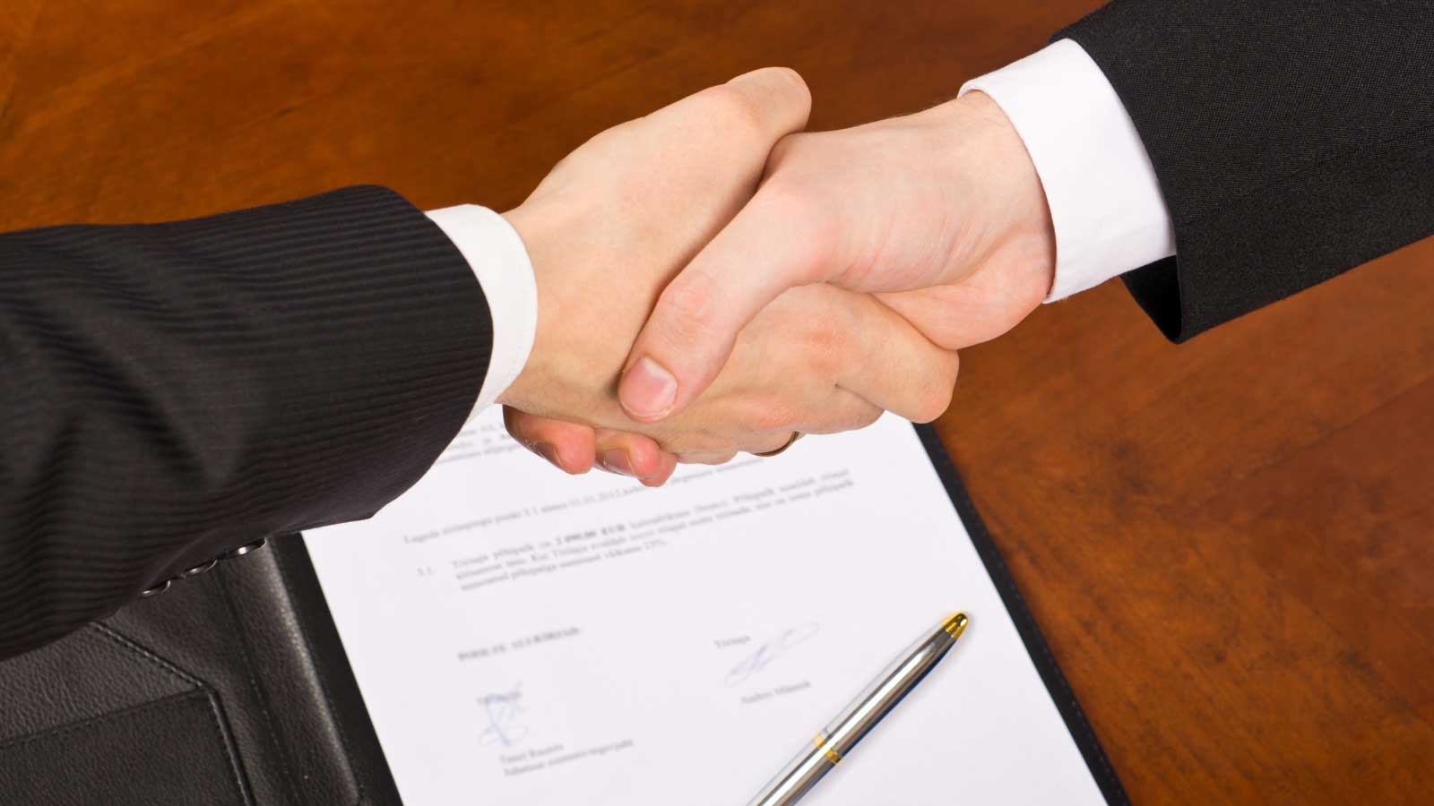 Two people in suits shaking hands over a signed contract on a wooden table