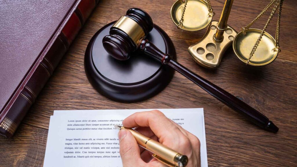 A hand holding a pen over a legal document on a desk with a gavel