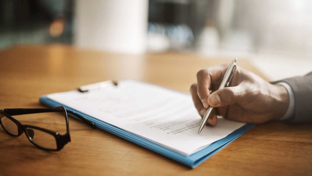 A person signs documents on a clipboard beside eyeglasses