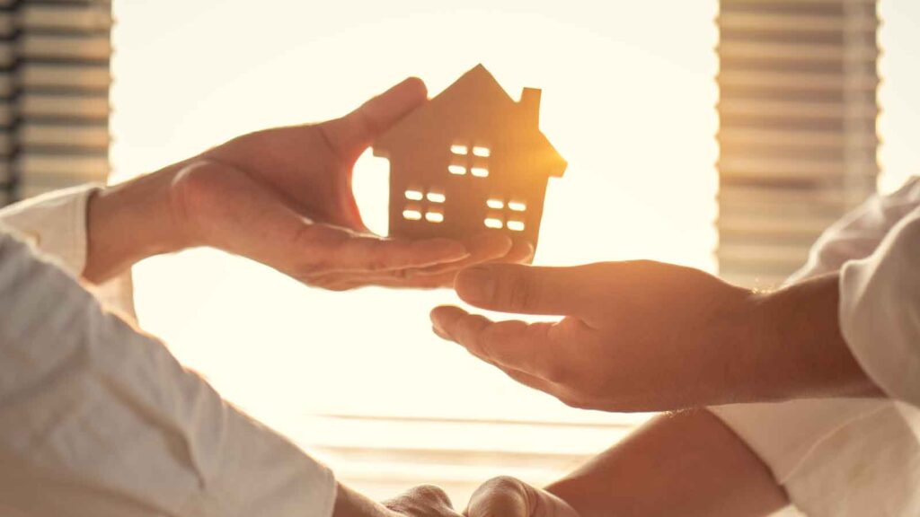 Two hands exchanging a small house model in warm sunlight.